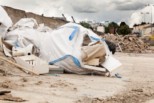 A large pile of discarded construction debris and waste materials located on an open, gravel-covered area. The pile includes white and beige waste bags partially torn open, revealing contents such as plastic pipes, broken fixtures, and packaging materials. Some items are strewn across the ground around the main heap, indicating recent disturbance or removal activity. In the background, there is a partially constructed or demolished wall, alongside scattered rubble and loose stones. Further in the distance, urban elements including buildings, streetlights, and a cloudy sky are visible, suggesting a typical urban environment that could be in Ealing or nearby areas. The scene is typical of rubbish removal or site clearance operations conducted by Commercial Waste Ealing, which specializes in local waste management services. The overall setting appears dusty and unmanaged, emphasizing the need for professional rubbish removal service to clear the site efficiently.