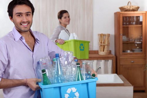 A smiling man with short dark hair and a light purple shirt holds a blue recycling bin filled with clear plastic bottles and containers, positioned in a kitchen with wooden cabinetry and a white sink. Behind him, a woman with dark hair pulled back, wearing a light grey top, stands near a green recycling bin labeled with a recycling symbol, on a wooden countertop alongside a tall, glass-fronted wooden cabinet. The kitchen background features a beige wall, a wicker basket on top of the cabinet, and natural lighting. The scene depicts household waste separation in a modern, tidy domestic setting, relevant to rubbish removal services like those provided by Commercial Waste Ealing in London, close to the Ealing area postcode.