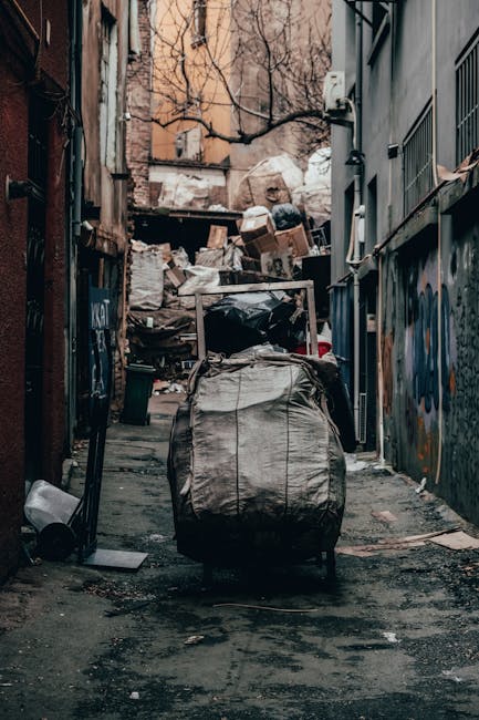 A narrow urban alleyway with uneven, dark asphalt ground, filled with significant amounts of accumulated waste and debris. At the forefront, a large, weathered grey plastic trash bag, partially open,