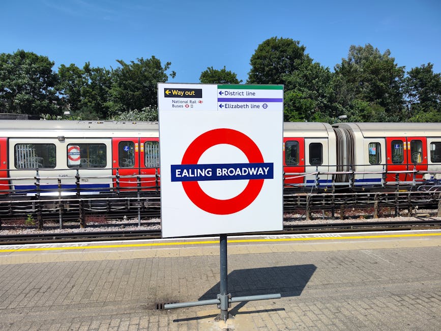 A close-up view of a white railway station sign displaying the name 'Ealing Broadway' in bold white letters on a blue background, mounted on a metal pole on a paved platform surface. Behind the sign, 